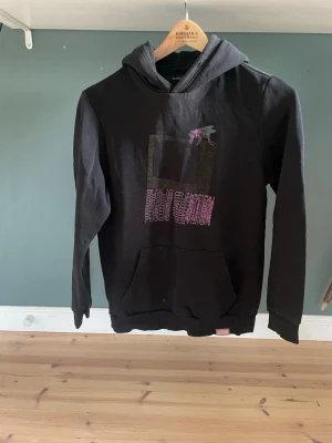Bershka Svart hoodie med tryck V.O.T.F - Svart hoodie från V.O.T.F med stor huva och magficka. Framsidan har grafiskt tryck i rosa och lila, baksidan har texten 'Visions of the Future' i flera rader och grafiska detaljer. Perfekt för dig som gillar streetwear och unika prints.