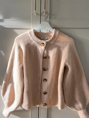 Beige/ rosa kofta - Mysig beige kofta från Indiska i ribbstickat material. Koftan har rund halsringning, stora bruna knappar och ballongärmar. Perfekt för lager-på-lager och enkel att matcha med olika outfits.