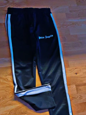 Palm Angels svarta mjukisbyxor - Svarta mjukisbyxor från Palm Angels med vita sidoränder och logga på vänster ben. Byxorna har resår i midjan och dragkedja vid benslut. Snygg streetwear-stil och skön passform, perfekta för en avslappnad look. Helt ny dress köpt för 1400  tröja 500 mjukis 400 eller hela dressen 700