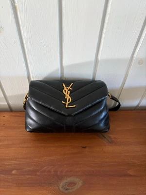 Svart YSL handväska i läder - Säljer en svart handväska från Saint Laurent med klassisk YSL-logga i guld framtill. Väskan är tillverkad i quiltat läder och har en axelrem i matchande svart med gulddetaljer. Perfekt storlek för det viktigaste och riktigt snygg design.