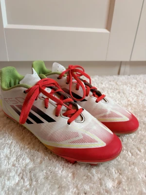 Adidas F50 vita och röda fotbollsskor - Säljer ett par Adidas F50 fotbollsskor i vitt med röda och svarta detaljer samt röda skosnören. Skorna har grönt foder och fasta dobbar i plast, perfekta för spel på gräs. 