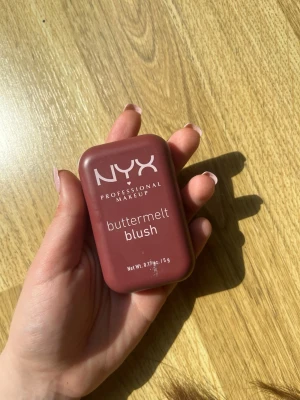 NYX buttermelt blush i rosabrun nyans - NYX Professional Makeup buttermelt blush i en snygg rosabrun nyans. Kommer i en kompakt, rektangulär dosa med spegel och har ett pressat mönster i pudret. Perfekt för att ge kinderna en naturlig och fräsch färg.