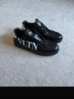 Svarta VLTN sneakers från Valentino - Snygga svarta sneakers från Valentino med VLTN-logga i vitt på sidan. Skorna är låga och har svarta skosnören samt vit detalj vid hälen. Tillverkade i läder med guldfärgad logga på plösen. Perfekta för dig som gillar exklusiva och trendiga sneakers.