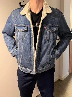 Levi's blå fodrad jeansjacka - Klassisk blå jeansjacka från Levi's med vit teddyfoder och krage. Jackan har två bröstfickor med lock och knapp, samt sidofickor. Ikonisk röd Levi's-tagg på bröstet och snygga söm-detaljer. Perfekt för dig som vill ha en tidlös och trendig look.