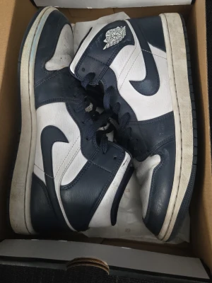 Nike Air Jordan 1 Mid Navy/Vit - Snygga Nike Air Jordan 1 Mid sneakers i marinblått och vitt läder. Klassisk high-top design med ikoniska Swoosh-loggan på sidan och Air Jordan Wings-logga vid ankeln. Perfekta för dig som gillar streetwear och vill sticka ut med en tidlös modell. Skriv för mer info