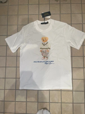 Vit Polo Bear t-shirt från Ralph Lauren - Vit t-shirt från Polo Ralph Lauren med ikoniskt Polo Bear-motiv på bröstet. Klassisk passform, rund halsringning och tryckt logga under björnen. Tillverkad i mjuk bomull för en skön känsla. Perfekt för en stilren och trendig look.