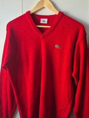 Vintage Lacoste v-neck - Röd stickad vintage tröja från Lacoste med klassisk V-neck och det ikoniska krokodilmärket broderat på bröstet. Tröjan har långa ärmar och ribbade muddar. Perfekt för dig som gillar stilrena och sportiga plagg.