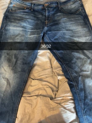 Blå jeans från Jack & Jones 36/32 - Säljer ett par klassiska blå jeans från Jack & Jones i storlek 36/32. Jeansen har en snygg tvätt med slitningar och kontrastsömmar. Tillverkade i bomull med fem fickor och normal passform. Perfekta för en avslappnad och trendig stil.