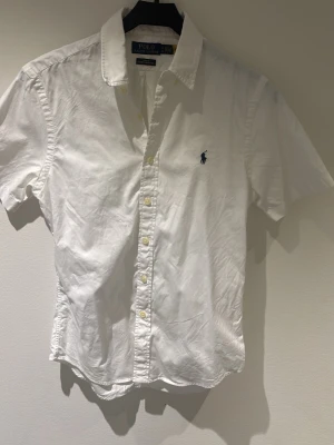 Vit kortärmad skjorta Polo Ralph Lauren - Vit kortärmad skjorta från Polo Ralph Lauren med blå logga på bröstet. Slim fit-modell i lätt bomullstyg, klassisk krage och knäppning framtill. Perfekt för en stilren och fräsch look. Storlek small köpt på Zalando. Använd extremt lite med perfekt skick. Hör av dig vid minsta fundering!