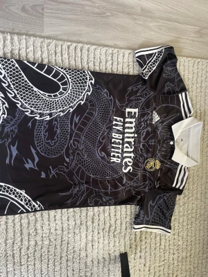 Real Madrid x Adidas svart fotbollströja - Snygg svart Real Madrid fotbollströja från Adidas med drakmönster i grått och vitt. Tröjan har vit krage, vita ränder på axlarna och klubbmärke broderat på bröstet. Perfekt för fotbollsträning eller att bära som supporter. PRISET KAN DISKUTERAS!