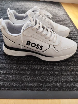 Vita sneakers från BOSS - Snygga vita sneakers från BOSS med svart logga på sidan. Skorna har en modern design med mesh och syntetmaterial, vita snören och svarta detaljer på sulan. Perfekta för dig som gillar stilrena och trendiga sneakers.