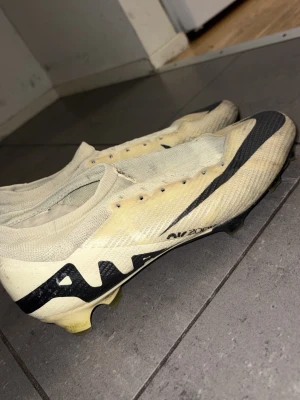 Nike Air Zoom Mercurial fotbollsskor - Säljer ett par Nike Air Zoom Mercurial fotbollsskor pros i beige med svarta detaljer och Nike-logga. Skorna har låg profil, snörning och ovandel i syntetmaterial för bra bollkontroll. Perfekta för dig som vill ha lätta och snabba skor på planen.