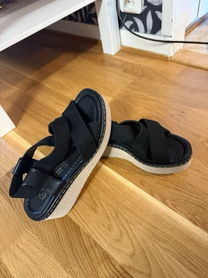 Svarta platåsandaler med korsade band - Säljer ett par svarta sandaler med breda korsade band i textil och läderimitation. Sandalerna har en beige platåsula med vit söm runt kanten och en mjuk innersula för extra komfort. Perfekta för dig som vill ha både stil och höjd. Den passar även storlek 38!  Helt ny och oanvänd!