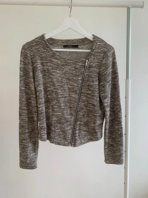 Gråmelerad cardiganjacka med dragkedja - Snygg gråmelerad cardiganjacka från Lindex med asymmetrisk dragkedja framtill. Jackan har rund halsringning, lång ärm och är stickad i ett mjukt material. Perfekt för lager-på-lager och enkel att matcha med olika outfits.