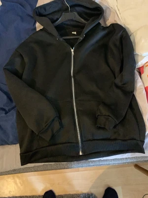 Svart zip-hoodie med huva - En klassisk svart hoodie med dragkedja framtill och stor huva. Tröjan har ribbade muddar vid ärmslut och nederkant samt två fickor framtill. Perfekt för en avslappnad och stilren look.