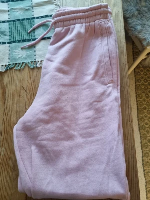 Ljusrosa mjukisbyxor med dragsko - Säljer ett par ljusrosa mjukisbyxor med resår och dragsko i midjan. Byxorna har raka ben och sidofickor. Perfekta för en avslappnad och chill stil. Materialet är mjukt och skönt, troligtvis bomull eller bomullsblandning.