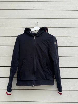 Svart hoodie från Moncler med dragkedja - Snygg svart hoodie från Moncler med dragkedja framtill, huva och kängurufickor. Klassisk Moncler-logga på vänster ärm och coola detaljer i rött och vitt vid ärmsluten. Perfekt för en stilren och avslappnad look. Nfc funkar. Bara ställ frågor om ni är intresserade!Är öppen för byten ochså! 