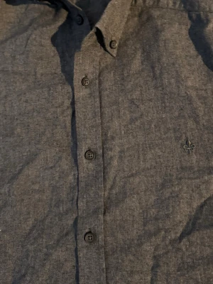 Grå skjorta från Morris🙌 - Säljer en grå skjorta med klassisk krage och knappar framtill. Skjortan har en diskret broderad detalj på bröstet och är tillverkad i ett mjukt bomullstyg. Perfekt för dig som gillar stilrena plagg med subtila detaljer.