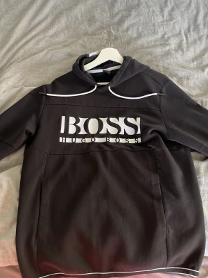 Svart hoodie från Hugo Boss - Snygg svart hoodie från Hugo Boss med stor vit logga på bröstet och vita detaljer längs huvan och nederkanten. Hoodien har en klassisk passform, stor ficka framtill och är tillverkad i mjukt bomullsmaterial. Perfekt för en avslappnad och trendig look.