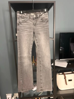 Grå bootcut jeans med broderade fickor - Snygga grå bootcut jeans med slitningseffekt och broderade bakfickor. Jeansen har klassisk femficksdesign, knappgylf och raka ben med lätt utsvängning. Perfekta för en trendig och avslappnad look.