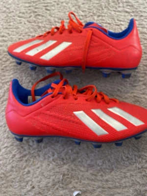 Röda Adidas X fotbollsskor - Säljer ett par röda Adidas X fotbollsskor med vita ränder och blå detaljer. Skorna har snörning och är tillverkade i syntetmaterial med dobbar för spel på gräs. Perfekta för dig som vill sticka ut på planen!