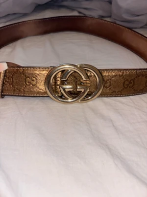 Gucci bälte i guld med GG-spänne - Snyggt bälte från Gucci i metallic guld med klassiskt GG-mönster och stort dubbelt G-spänne i guld. Bältet är gjort i skinn och har en lyxig känsla som verkligen sticker ut.