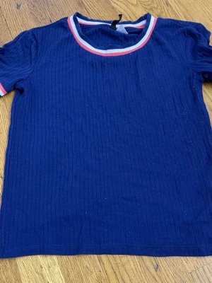 Marinblå ribbad t-shirt med ränder - Snygg marinblå ribbad t-shirt med korta ärmar. Kragen och ärmsluten har vita och rosa ränder som ger en sportig vibe. Perfekt till jeans eller kjol för en avslappnad look.