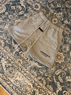 Grå Essentials shorts från Fear of God - Säljer ett par grå mjukisshorts från Essentials Fear of God. De har elastisk midja med snörning, sidofickor och logga på vänster ben. Perfekta för chill dagar eller träning. Tillverkade i mjukt bomullsmaterial för maximal komfort.