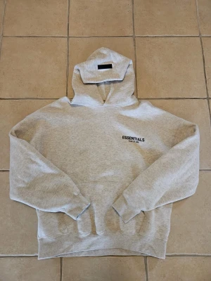 Grå Essentials Fear of God hoodie - Snygg ljusgrå hoodie från Fear of God Essentials med svart logga på bröstet och stor text på ryggen. Klassisk passform med huva och ribbade muddar. Perfekt för en avslappnad streetwear-look.