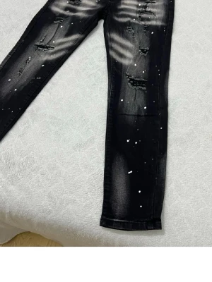 Svarta slitna jeans med färgstänk - Säljer ett par svarta jeans med slitningar och coola vita färgstänk. Jeansen har skinny fit och tydliga slitdetaljer på låren och knäna. Perfekta för dig som gillar en edgy streetstil. Materialet är klassisk denim med stretch.