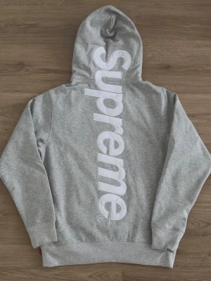 Grå Supreme hoodie med stor logga - Supersnygg grå hoodie från Supreme med stor vit logga längs ryggen. Klassisk känguruficka framtill, huva med snörning och ribbade muddar. Perfekt för dig som gillar streetwear och vill sticka ut med en ikonisk look.