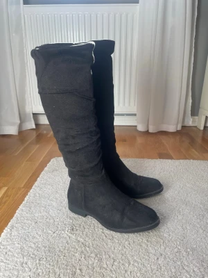 Svarta höga mockastövlar - Snygga svarta stövlar i mocka med låg klack och rundad tå. Stövlarna har en slouchy passform och är perfekta för att lyfta vilken outfit som helst. Klassisk och stilren design som funkar till många looks.