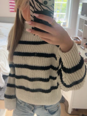 Randig stickad tröja från H&M - Säljer en krämvit stickad tröja från H&M med svarta horisontella ränder. Tröjan har en avslappnad passform, ribbstickade muddar och hög hals. Perfekt för att hålla sig varm och snygg under kyliga dagar.