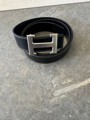Svart skärp med H-spänne - Stilrent svart bälte med ett stort, silverfärgat H-format spänne. Bältet har vita kontrastsömmar längs kanterna och är gjort i skinn. Perfekt accessoar för att lyfta din outfit.