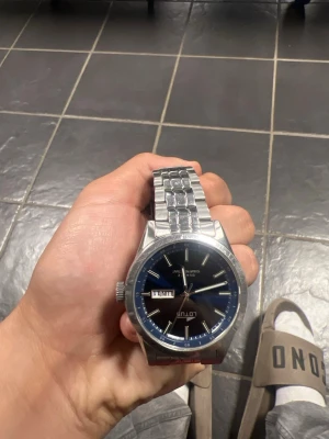 Silverfärgad Lotus herrklocka - Stilren herrklocka från Lotus med silverfärgat armband i metall och svart urtavla. Klockan har dag- och datumvisning samt tydliga indexmarkeringar. Perfekt accessoar för dig som gillar klassisk design.