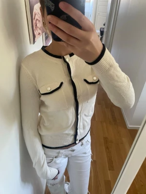 Vit kofta med svarta detaljer - Snygg vit kofta med svarta kanter och dekorativa fickor framtill. Rund halsringning och knäppning hela vägen. Perfekt för en stilren look och enkel att matcha med andra plagg.