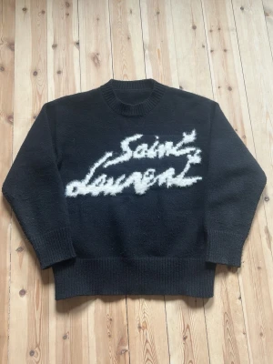 Svart Saint Laurent stickad tröja - Cool svart stickad tröja från Saint Laurent med vit fuzzy logotyp framtill. Tröjan har rund halsringning, ribbade muddar och är tillverkad i Italien. Perfekt för dig som gillar exklusiva och trendiga plagg.