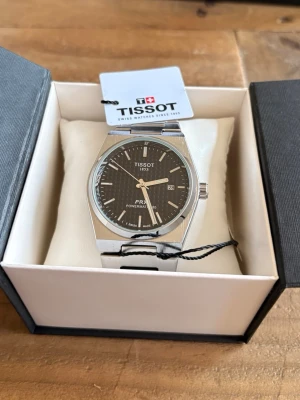 Tissot PRX Powermatic 80 klocka silver - Snygg Tissot PRX Powermatic 80 klocka i silverfärgat stål med svart urtavla och datumvisning. Klockan har en stilren, modern design med länkarmband och tydliga indexmarkeringar. Perfekt accessoar för dig som gillar klassisk men trendig stil.
