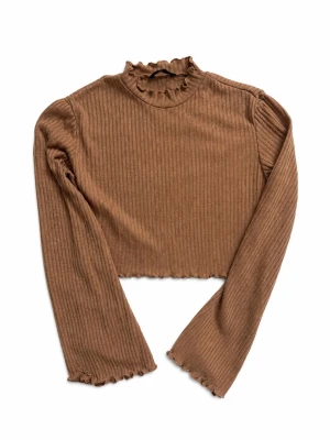 Brun ribbad croppad tröja - Trendig brun långärmad tröja med ribbad struktur och croppad passform. Hög hals med volangkant och volangdetaljer vid ärmslut och nederkant. Perfekt för lager-på-lager och snygg till jeans eller kjol.