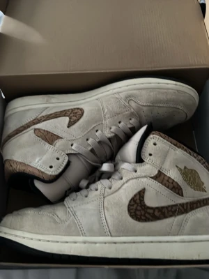 Air Jordan 1 Mid SE Orewood Brun - Snygga Air Jordan 1 Mid SE i färgen Orewood Brun med metallicguld-detaljer. Skorna har en beige mockaöverdel, bruna swoosh-loggor med mönster och klassisk Jordan Wings-logga i guld på sidan. Perfekta sneakers för dig som gillar unik design och streetwear.