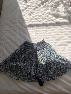 Blommig mini kjol med volang💙 - Supersöt kjol med blått och vitt blommönster och volangkant. Materialet känns lätt och luftigt, och midjan har en dekorativ knytning framtill. Jag har sytt på sidan om höfterna för att den annars vart lite för stor för mig i höfterna. Midija ca: 56cm (det går att sy upp det jag sytt och få en större midja). längd från midja: ca 33cm