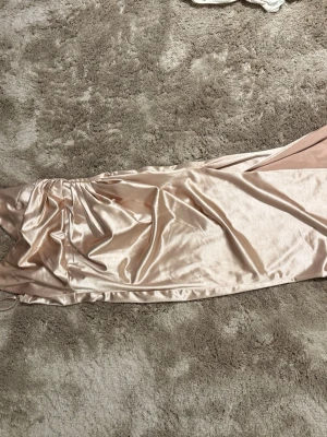 Champagnefärgad långklänning i satin - Elegant långklänning i skimrande champagnefärgad satin med figurnära passform och en öppning i ena benet. Klänningen har smala axelband och en draperad detalj vid sidan. Obs: kommer från shein, använd 1 gång på min bal i 9:an, sytt på den själv (syns inte). Säljs pga: använder aldrig 