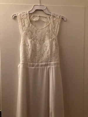 Vit spetsklänning med öppen rygg - Elegant vit klänning med spets upptill och öppen ryggdetalj. Klänningen har en mjuk, flowy kjol i slätt tyg. Spetsen har blommigt mönster och ger en romantisk känsla. Perfekt för dig som vill ha en stilren och feminin look.