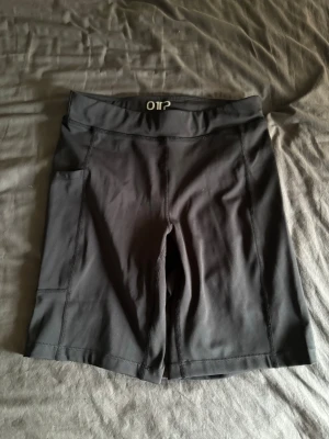 Svarta träningstights shorts OTP - Svarta träningstights shorts från OTP med hög midja och praktisk ficka på sidan. Tillverkade i stretchigt syntetmaterial som passar perfekt för aktiva dagar. Snygg och enkel design som är lätt att matcha med andra träningskläder.