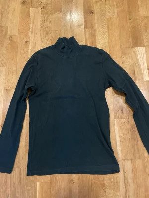 Samsöe grön polo  med lång ärm stl M  - En grön polo med lång ärm och klassisk passform. Toppen har en hög krage och är tillverkad i ett mjukt material som känns skönt mot huden. Perfekt att styla lager på lager eller bära som den är.