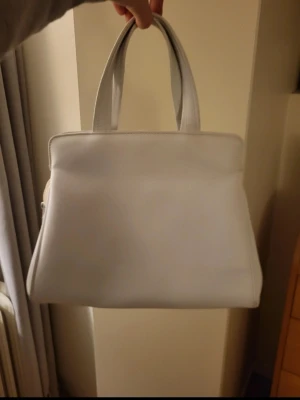 Vit handväska med guldiga detaljer - Vit handväska i stilren, fyrkantig modell med två stadiga handtag. Väskan har snygga guldiga metalldetaljer vid handtagen och är gjord i ett material som ser ut som läder. Perfekt accessoar för dig som gillar minimalistisk och klassisk design.