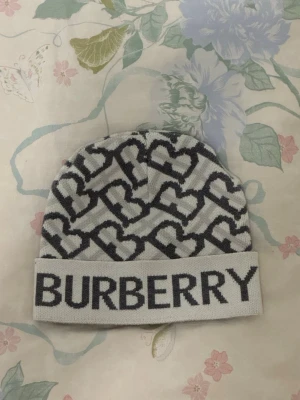 Burberry mössa  - Snygg stickad mössa från Burberry i ljusgrått med mörkgrå hjärtmönster och stor BURBERRY-logga framtill. Mössan har uppvikt kant och är tillverkad i mjukt stickat material som håller dig varm och trendig.