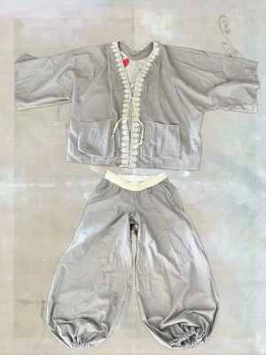 Grått set med vida byxor och kimono - Unikt grått tvådelat set med vida byxor och matchande kimono. Kimonon har stora fickor och dekorativ spetskant samt knytning framtill. Byxorna har resår i midja och benslut för en avslappnad look. Perfekt för dig som gillar oversized och bohemisk stil. Passar M-L då byxorna har resår! 