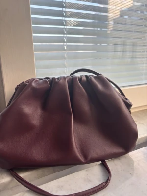 Vinröd clutchväska i skinnimitation - Trendig vinröd handväska med mjuk, rynkad design och dragskostängning. Väskan har en slät yta i skinnimitation och en smal axelrem. Perfekt accessoar för att lyfta din outfit.