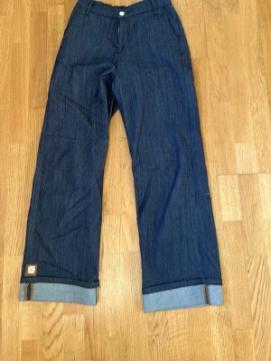 Blåa vida jeans, kostym byxor - Säljer ett par mörkblå jeans med bred passform och uppvikta benslut. Jeansen har klassisk femficksdesign, knappgylf och en liten patchdetalj vid benet. Perfekta för en avslappnad och trendig look.